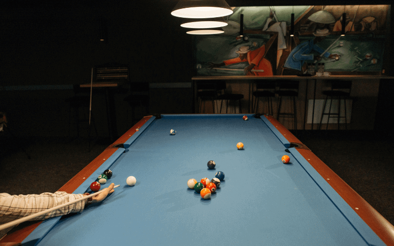 A hand holding a cue stick, applying maintenance tools to clean and polish the cue, with a rack of billiard balls in the background